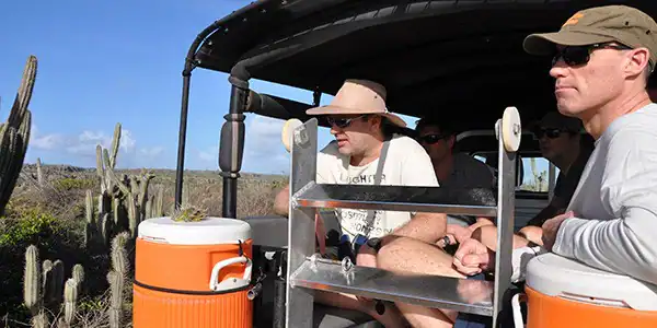 Cuatro personas disfrutan de un safari en Jeep al Parque Nacional Christoffel (Curazao) en un vehículo al aire libre, con sombreros y gafas de sol, mientras observan altos cactus y paisajes desérticos. En el jeep se pueden ver dos nevera naranja.