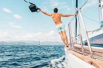Een man in zwembroek staat op de rand van een zeilboot, met een tas boven zijn hoofd, met blauw oceaanwater en zeilboten in de verte zichtbaar onder een gedeeltelijk bewolkte hemel.