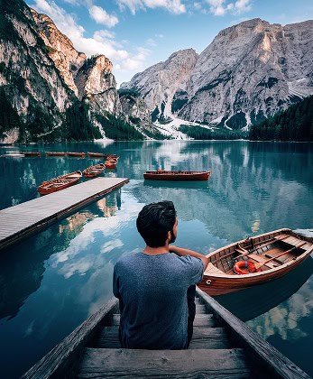 Een persoon zit op een houten trapje bij een rustig alpenmeer met houten bootjes die in de buurt dobberen. Majestueuze bergen en dennenbomen omringen het meer onder een gedeeltelijk bewolkte hemel, wat een serene en schilderachtige sfeer creëert.