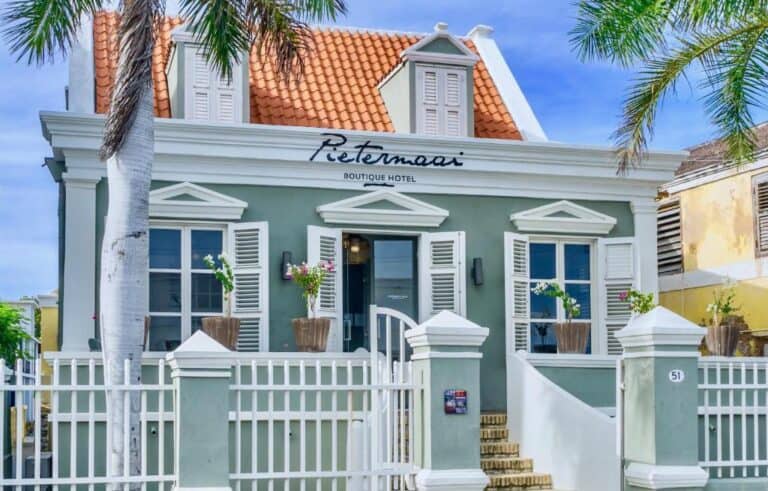 A small green boutique hotel with white shutters and a red tiled roof, named Pietermaai, behind a white fence, with potted plants at the entrance and palm trees in front.
