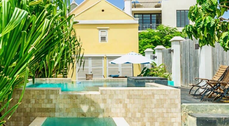 A small yellow house with white trim stands behind a stone-tiled pool, surrounded by lush green plants. Lounge chairs and a white umbrella sit on a stone patio under a bright, sunny sky.
