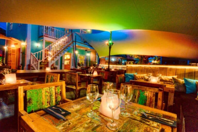 Cozy outdoor restaurant with rustic wooden tables, cushioned seating, and glowing candles. Colorful lights illuminate the space, and a staircase leads to an upper level, creating a warm and inviting atmosphere at night.