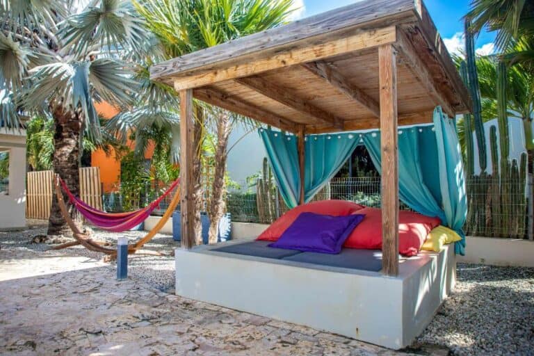 Een schaduwrijk buitenbed met kleurrijke kussens en groenblauwe gordijnen staat op een stenen patio tussen palmbomen, met een roze, gele en paarse hangmat in de buurt onder een zonnige hemel.