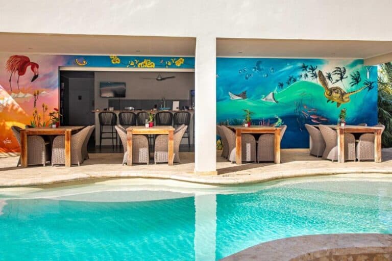 Een bar aan het zwembad met rieten stoelen en houten tafels tegen levendige muurschilderingen van een flamingo, een oceaanschilderij met schildpadden en zeeleven, die een tropische, ontspannen sfeer creëren.