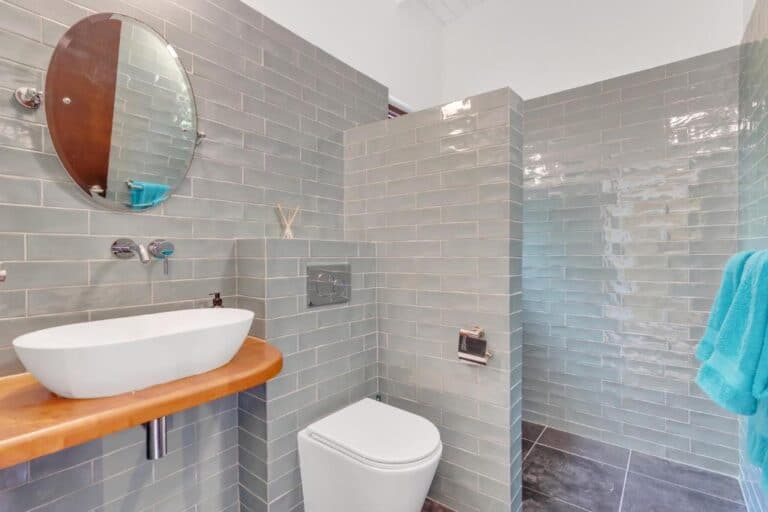 Moderne badkamer met lichtgrijze metro tegels, een ronde spiegel boven een witte wasbak op een houten aanrecht, een wandtoilet en een inloopdouche. Aan de muur hangt een blauwe handdoek.