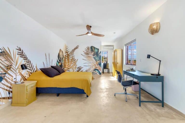 Ruime, moderne slaapkamer met een groot bed, geel beddengoed, muurkunst van tropische bladeren, een plafondventilator, geel nachtkastje, blauw bureau met lamp en stoel en grote ramen die natuurlijk licht binnenlaten.