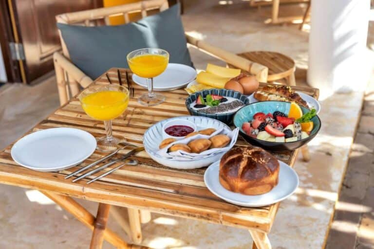 Een houten tafel gedekt voor het ontbijt met twee glazen sinaasappelsap, een bord fruit, brood, gebak, kaas, gekookte eieren en jam, met twee lege borden en keukengerei naast bamboe stoelen.