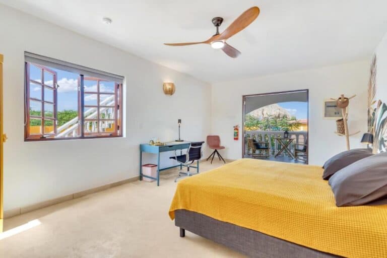 Lichte slaapkamer met een gele sprei, plafondventilator, bureau en stoel. Grote ramen en een glazen deur openen naar een balkon met zitje en een rieten dak, waardoor er veel natuurlijk licht binnenvalt.