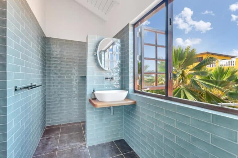 Een moderne badkamer met lichtblauwe metrotegels, een ronde spiegel, een witte wastafel op een houten aanrecht, een open raam met palmbomen en een blauwe lucht met wolken.