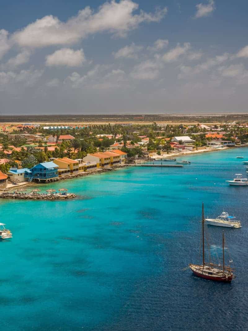 Luchtfoto van een kustplaats met levendig turquoise water, boten voor anker voor de kust, kleurrijke gebouwen en een gedeeltelijk bewolkte blauwe lucht.