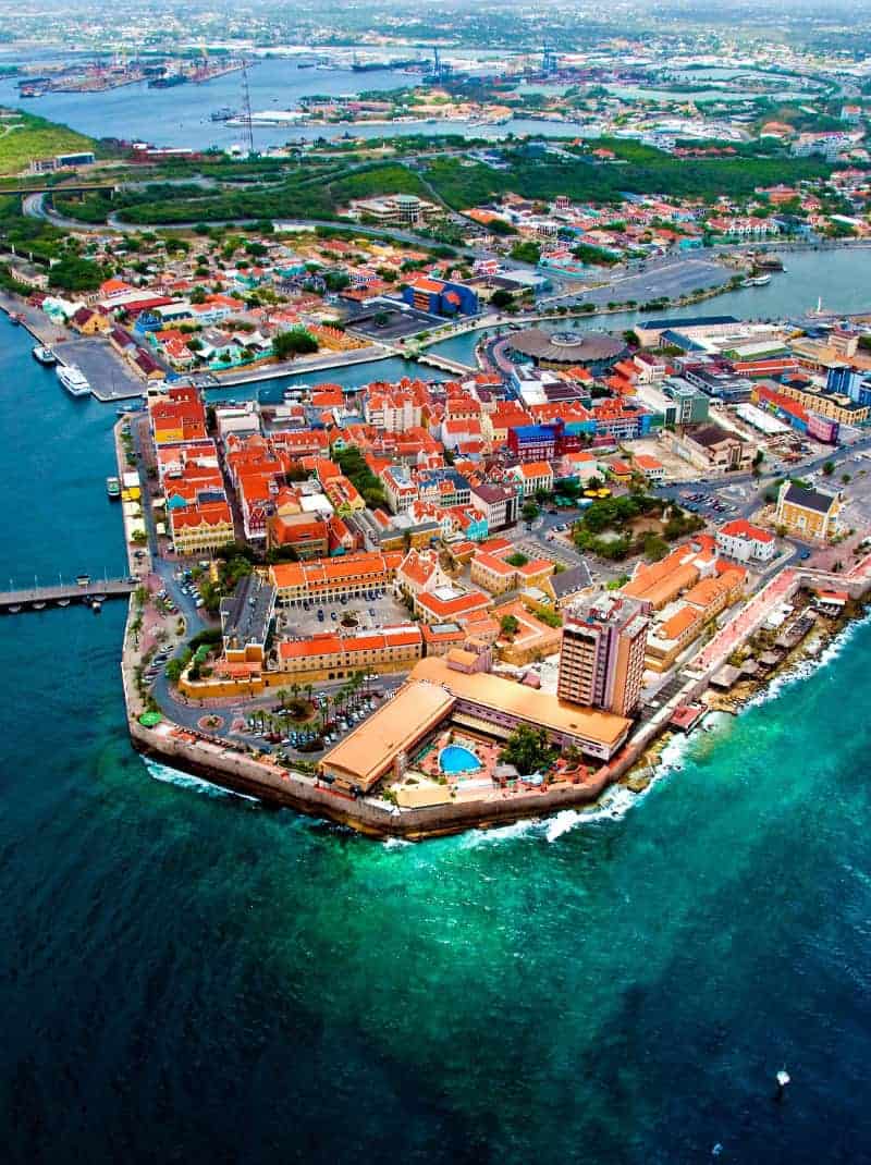 Luchtfoto van een kuststad met kleurrijke gebouwen, omringd door turquoise water, een haven en een mix van woonwijken en commerciële gebieden. De stad wordt begrensd door een zeewering en heeft een hoog hotel met een zwembad bij de kust.