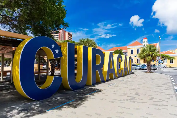 Cartel grande y colorido de CURACAO con estrellas, situado en una soleada plaza bordeada de palmeras y edificios históricos amarillos bajo un cielo azul brillante.