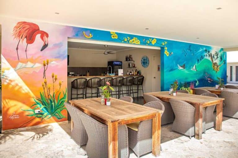 Helder, kleurrijk café met houten tafels en rieten stoelen, met levendige muurschilderingen op de muren van een flamingo, planten en oceaanscènes. De bar heeft zwarte krukken en achter de toonbank staan flessen uitgestald.