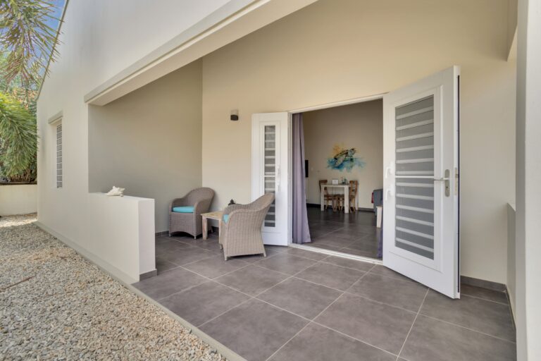 Een moderne patio met twee rieten stoelen en een kleine tafel op grijze tegels, die leidt naar dubbele glazen deuren die uitkomen in een lichte binnenkamer met kunstwerken aan de muur. De patio wordt begrensd door witte muren en grind.
