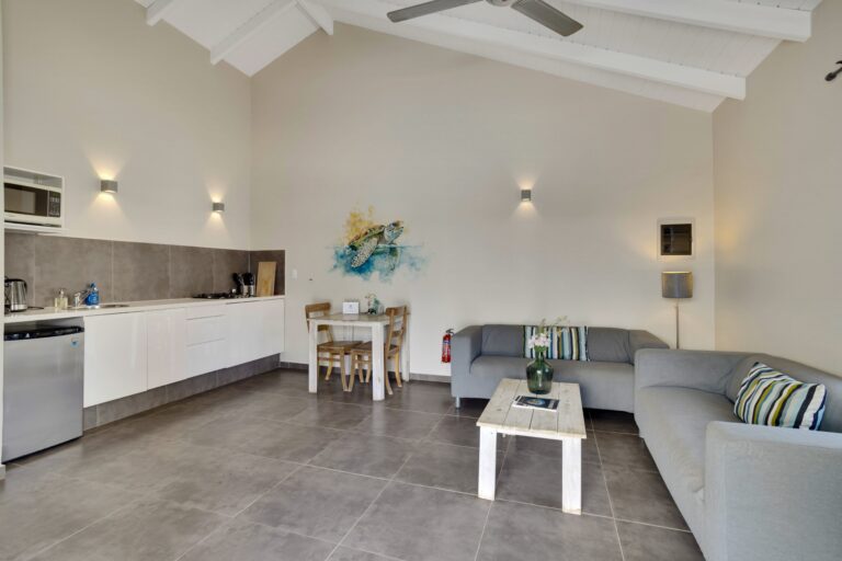 Moderne woonkamer met grijze tegelvloer, lichte banken, een witte salontafel, een kleine eettafel met stoelen, een kitchenette met apparatuur en muurkunst. Hoog plafond met balkenplafond en plafondventilator.