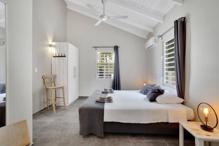Een moderne slaapkamer met een tweepersoonsbed, grijs beddengoed en kussens, twee bijzettafeltjes met lampen, een kledingkast, een houten stoel, een wandspiegel, grijze gordijnen en een plafondventilator onder een wit gewelfd plafond.