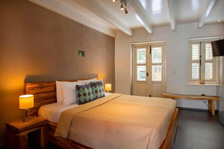 A cozy bedroom with a large wooden bed, beige bedding, two bedside tables with lamps, decorative pillows, a small bench under the window, and sunlight coming through shutters on double doors.