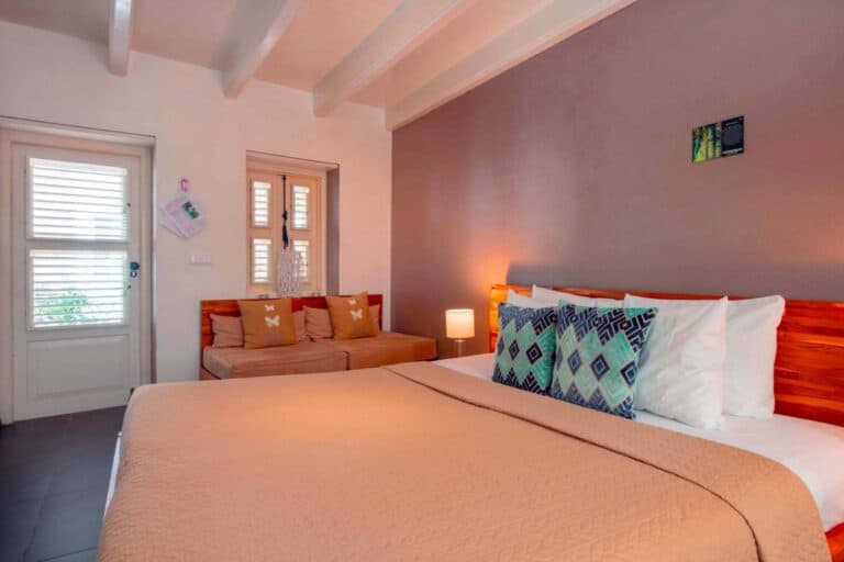 A cozy bedroom with a large bed, decorative pillows, a beige quilt, a wooden headboard, a cushioned bench with pillows, two small windows, a lamp, and soft lighting creating a warm atmosphere.