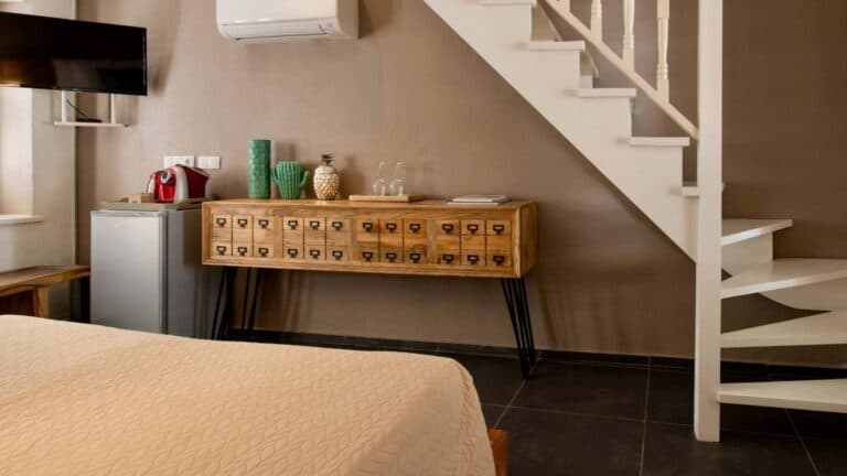 A cozy room with a beige bedspread, a small fridge, a coffee maker, decorative vases, and glasses on a wooden sideboard under a staircase with white steps and railing. A TV is mounted on the wall.