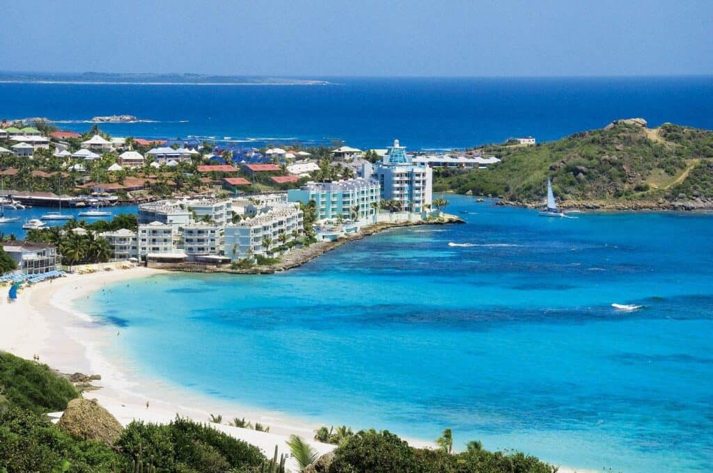 A coastal view of white sandy beaches, turquoise blue water, and a marina with buildings and resorts along the shoreline; a sailboat is seen in the bay, with green hills in the background.