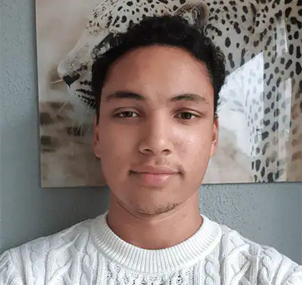 Een jong persoon met kort krullend haar en een witte kabeltrui staat voor een muur met een ingelijste foto van een luipaard op de achtergrond.