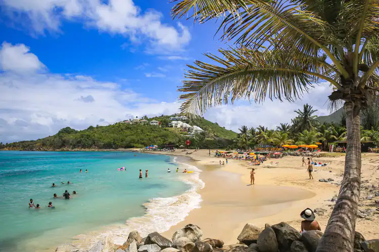 La gente nada y se relaja en una playa tropical de arena con aguas turquesas, palmeras y colinas a lo lejos. Paraguas coloridos y vegetación a lo largo de la costa bajo un cielo azul con nubes dispersas.