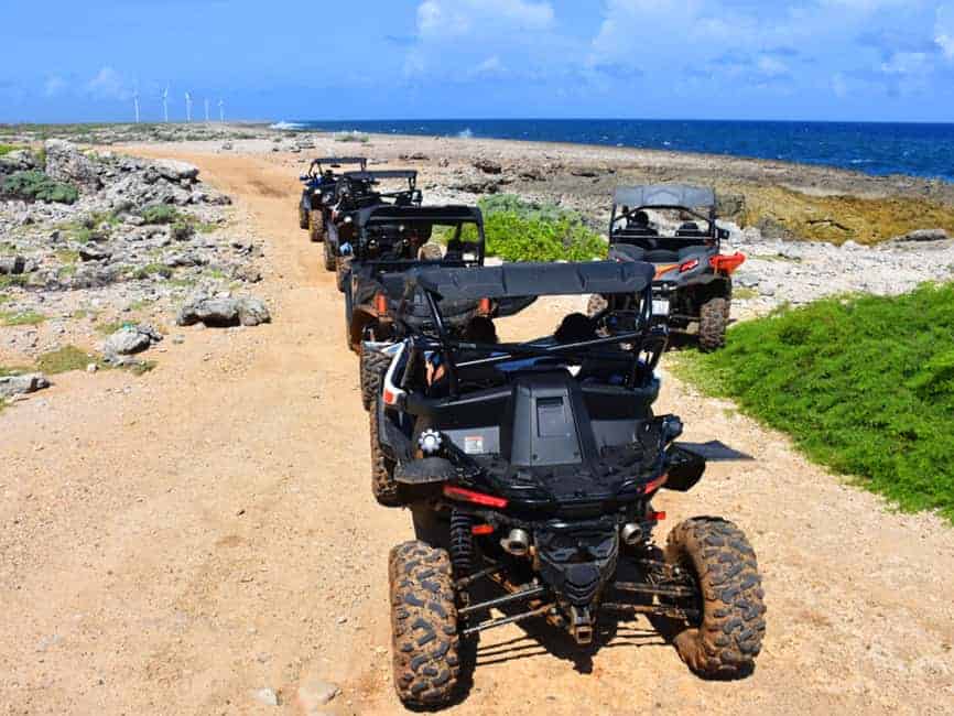 Una fila de buggies todoterreno del Off-Road Buggy Tour (Curazao) recorre un sendero costero rocoso, con el océano azul a la derecha y molinos de viento a lo lejos bajo un cielo parcialmente nublado.