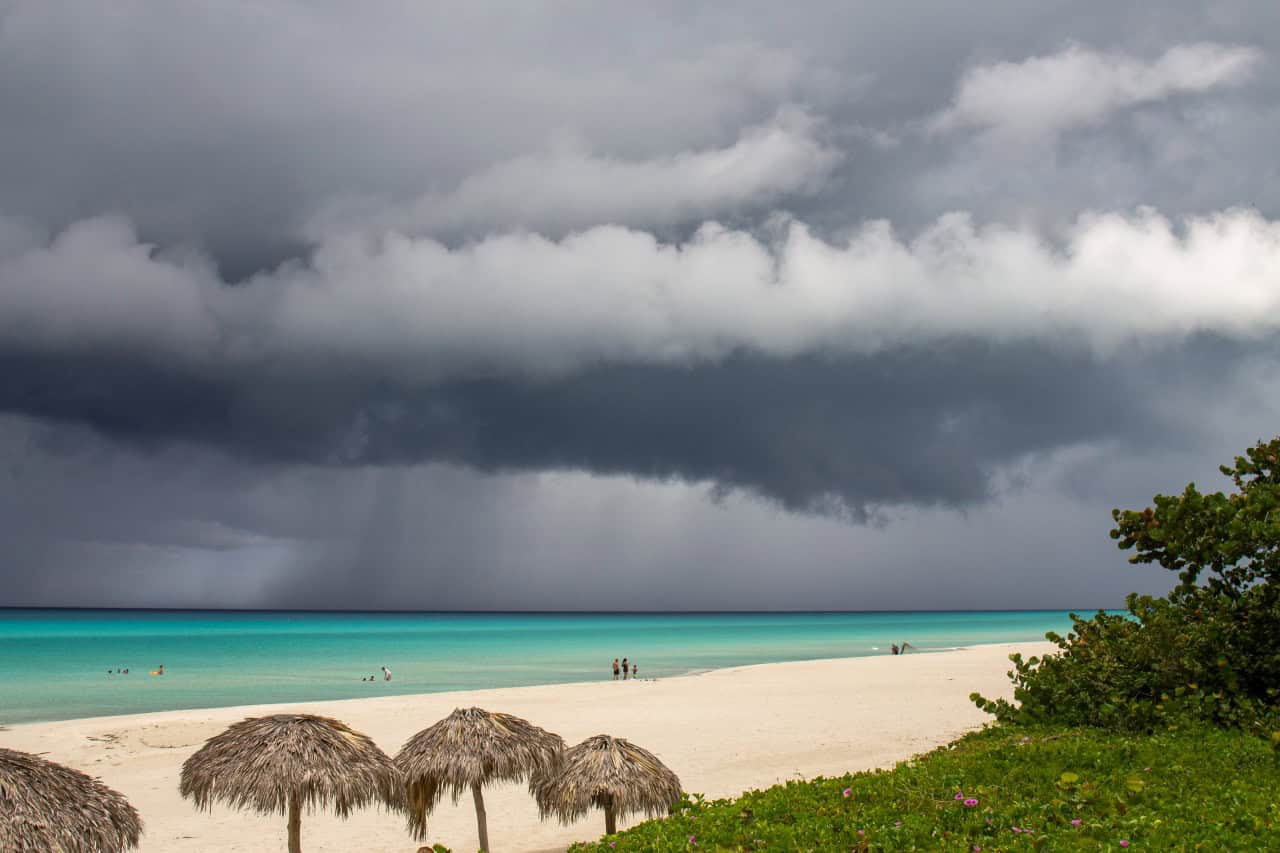 Een strand met wit zand, turquoise water en rieten parasols ligt onder een dramatische hemel terwijl donkere stormwolken naderen - typisch voor het regenseizoen op Bonaire. Een paar mensen wandelen langs de kustlijn, omzoomd door weelderige groene planten.