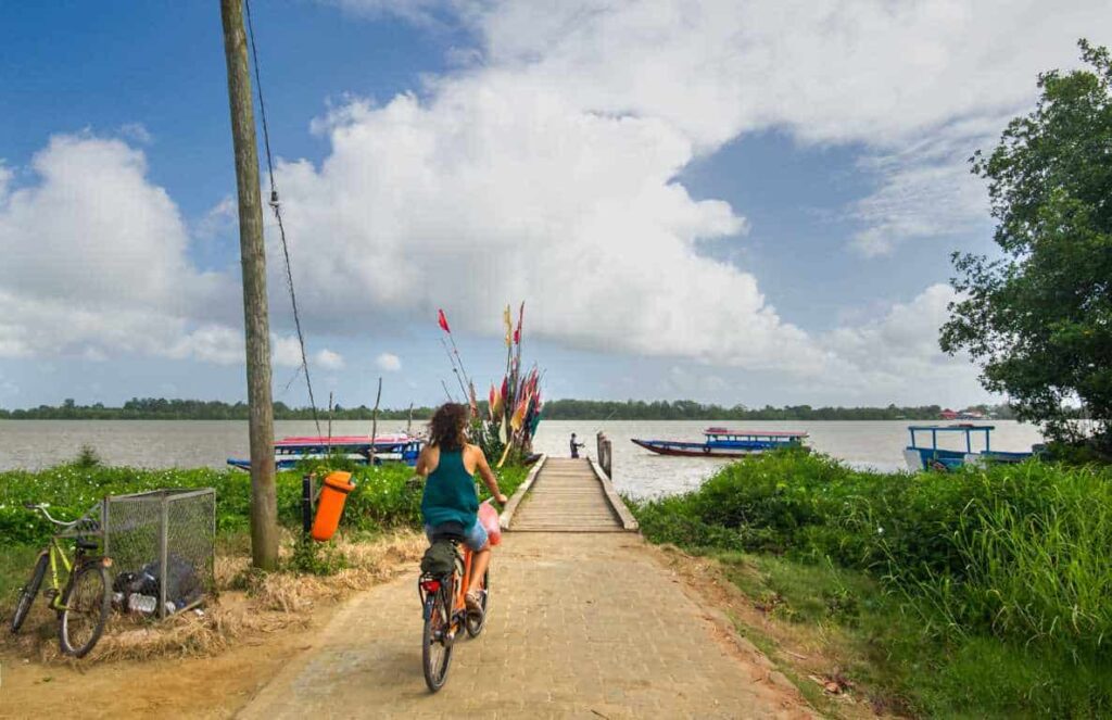 Een persoon fietst over een geplaveid pad naar een houten steiger met kleurrijke vlaggen bij de Peperpot Tour - Historische Plantage & Natuurpark (Suriname), langs weelderig groen gras en boten die langs de rivier liggen onder een heldere, bewolkte hemel.
