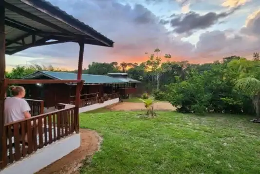 Een persoon op een houten veranda kijkt uit over een grasveld en palmbomen, met gebouwen met groene daken tussen de bomen onder een zonsondergang - een foto van de Suriname River Kayak Expeditie (Suriname).