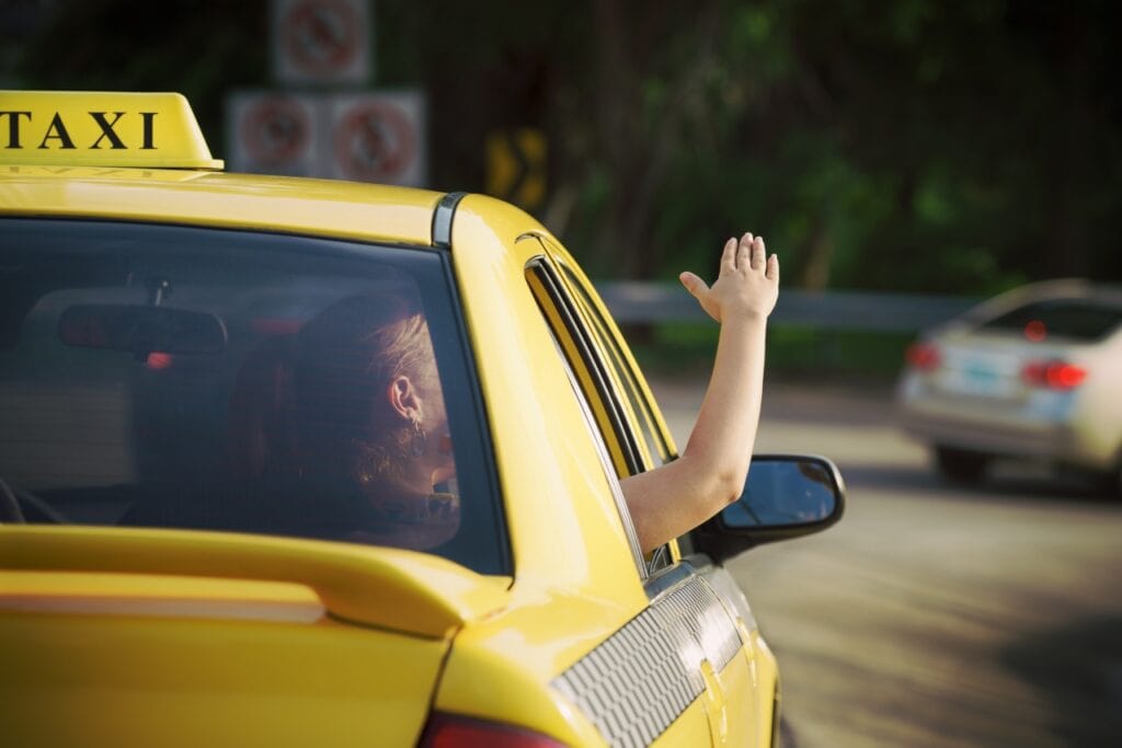 A person in the back seat of an airport transfer taxi in Suriname, leans out the window and waves his hand while the taxi is on a road in. Suriname drives with other cars and greenery in the background.