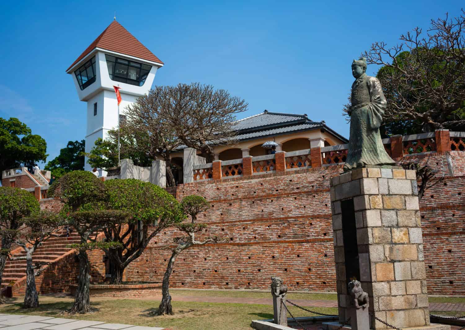 A statue of a historical figure stands on a stone pedestal in front of manicured trees and a red brick wall near Fort Zeelandia, with a white observation tower topped by a red roof under a clear blue sky in the background.
