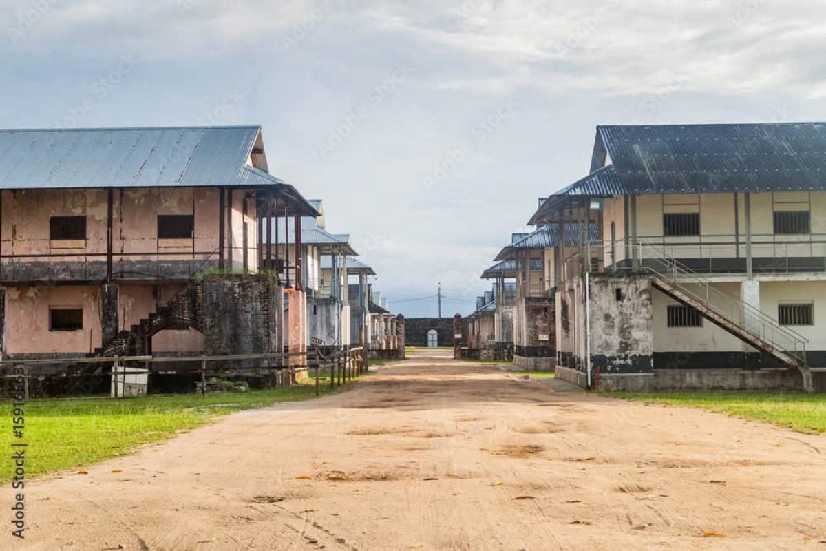 On the French Guiana Day Trip (Suriname), you will see two rows of old buildings with metal roofs and stairs along a wide sandy path, lined by green grass and under a cloudy sky.