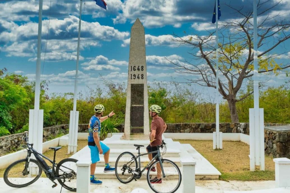 Twee fietsers met helmen staan bij hun fiets bij een monument, gemarkeerd 1648 en 1948, op de "Two Countries, One Bike Tour (Sint Maarten)", omringd door vlaggenmasten en bomen onder een gedeeltelijk bewolkte hemel.