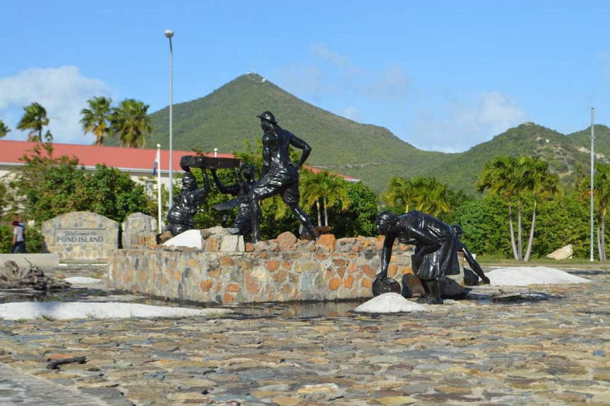 Experimente el "Recorrido Histórico en Bicicleta y Tiempo en la Playa (San Martín)": vea estatuas negras de trabajadores, exuberantes montañas verdes, palmeras y un edificio de tejados rojos bajo un cielo azul.