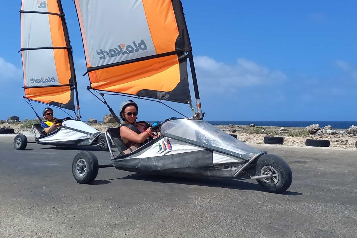 Twee personen genieten van een 30 minuten durende Landzeiltocht op Bonaire, waarbij landjachten met oranje zeilen over een geplaveid kustpad varen, omringd door rotsachtig terrein en oceaan onder een helderblauwe hemel.