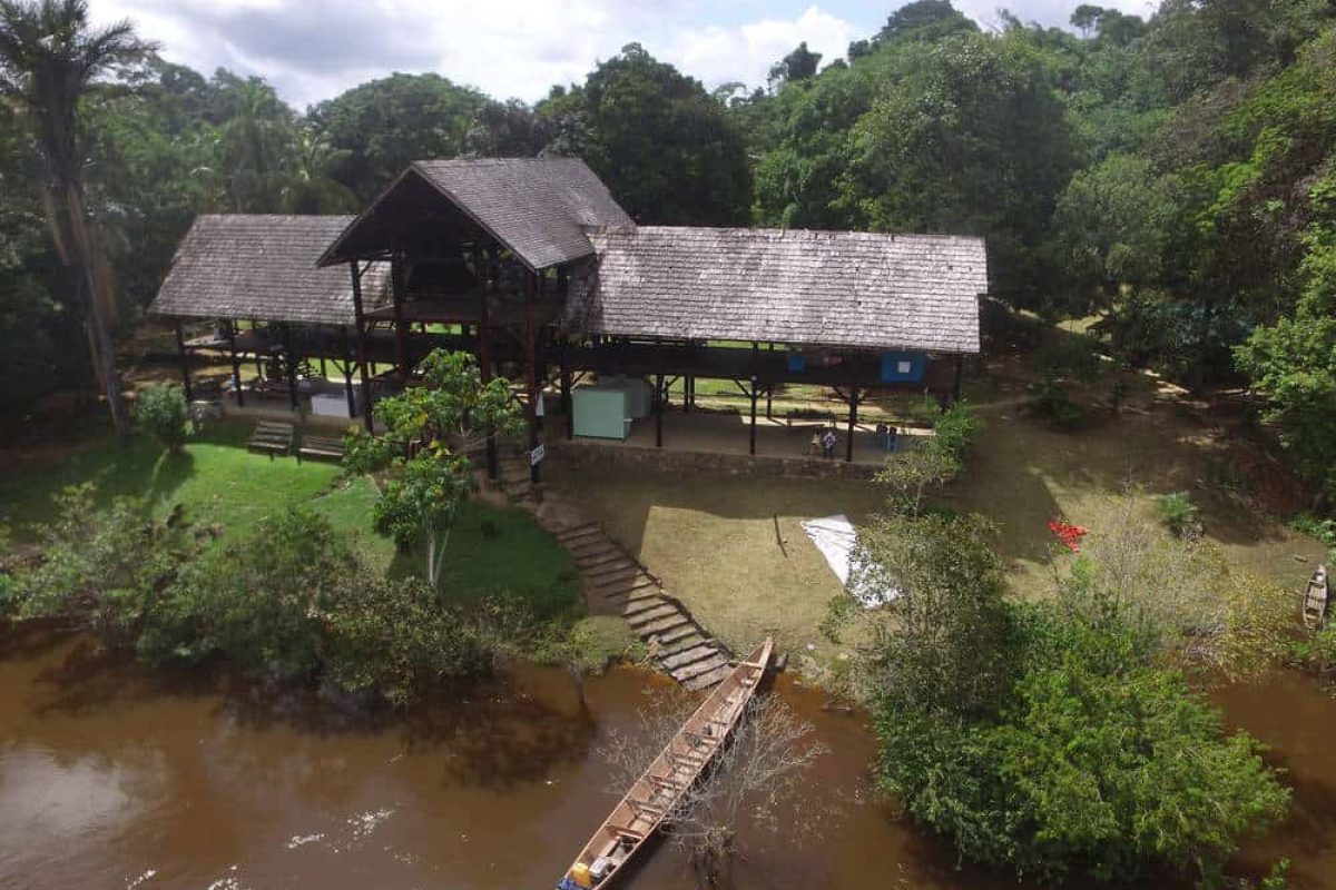 Ervaar de 4 Dagen Raleighvallen Voltzberg (Suriname): een houten lodge met een hoog dak, gelegen op palen aan een bruine rivier te midden van weelderig groen, met een kano aangemeerd bij een stenen pad dat naar de lodge leidt.