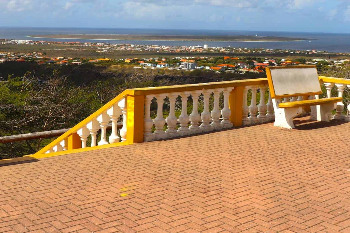 El Tour Privado del Norte (Bonaire) cuenta con un banco amarillo y blanco sobre una terraza de ladrillo con barandilla decorativa, con vistas pintorescas de la ciudad costera de Bonaire, la vegetación exuberante y el mar bajo un cielo parcialmente nublado.