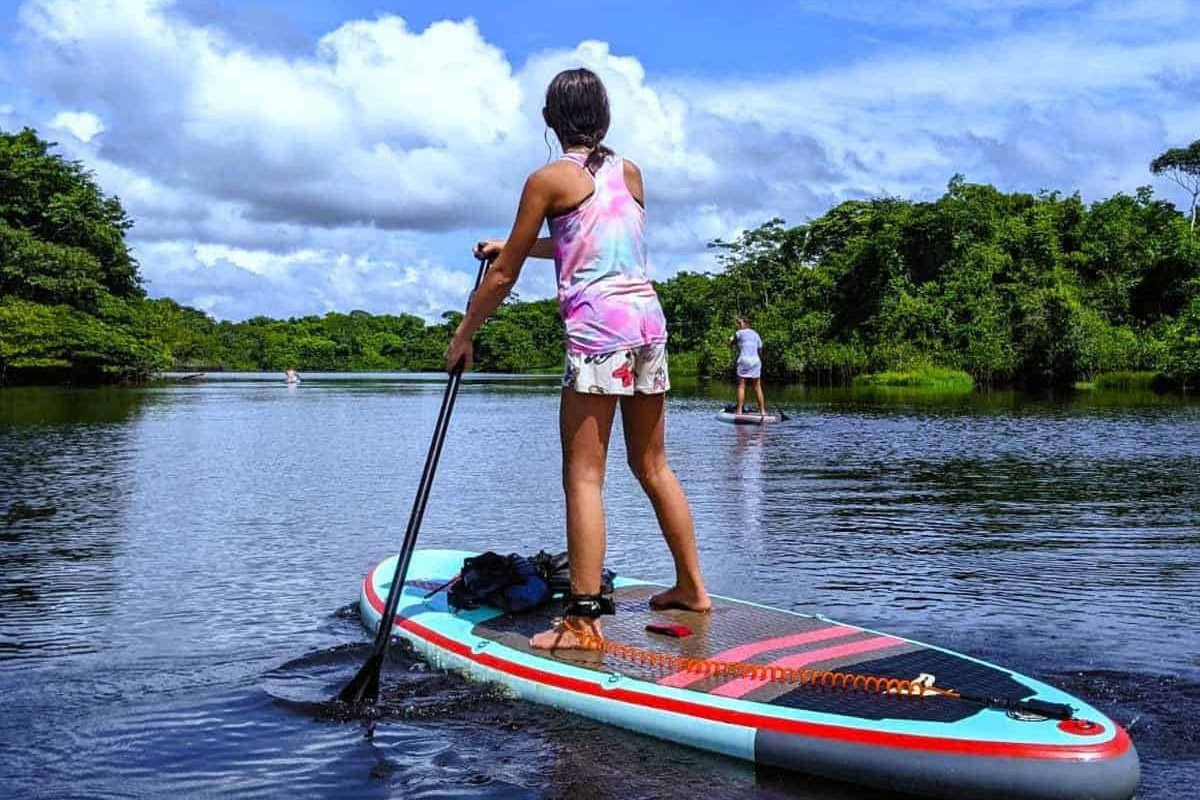 Een persoon staat op een paddleboard in kalm water met een peddel in de hand tijdens de Dagtocht Frederiksdorp (Suriname), met een andere peddelaar in de verte en weelderige groene bomen onder een gedeeltelijk bewolkte hemel.