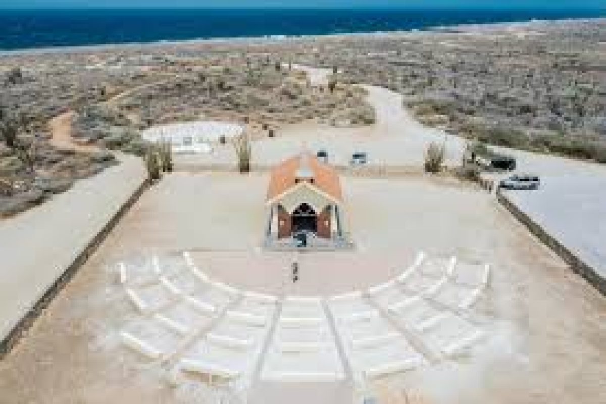 Experience the Mangel Halto & Baby Beach Off-Road Safari in Aruba, where a red-roofed chapel stands in an arid landscape, white benches in a semicircle, with winding roads and ocean views that complete the picturesque adventure.