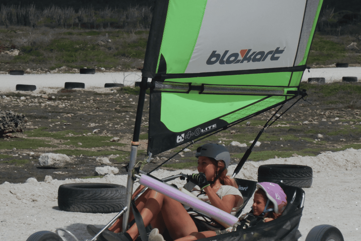 Een volwassene en kind rijden 30 minuten in een driewielige Landzeilen kart op Bonaire. Ze sturen samen onder een groot groen en grijs zeil op een zandpad. Beiden dragen een helm; op de achtergrond zijn banden te zien.