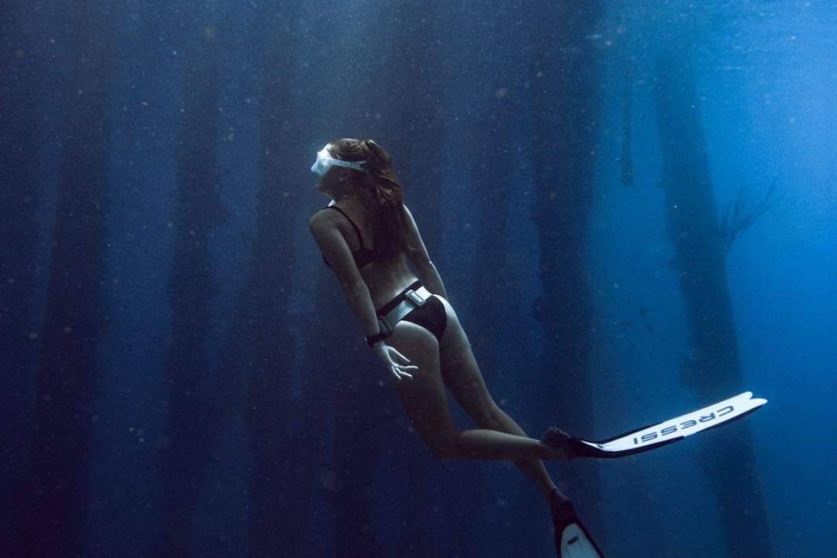 Una mujer con equipo de snorkel y aletas nada hacia la superficie en aguas azul profunda, con la luz del sol a su alrededor, durante la experiencia de Freediving Adventure Dive Reef Exploration (Bonaire).