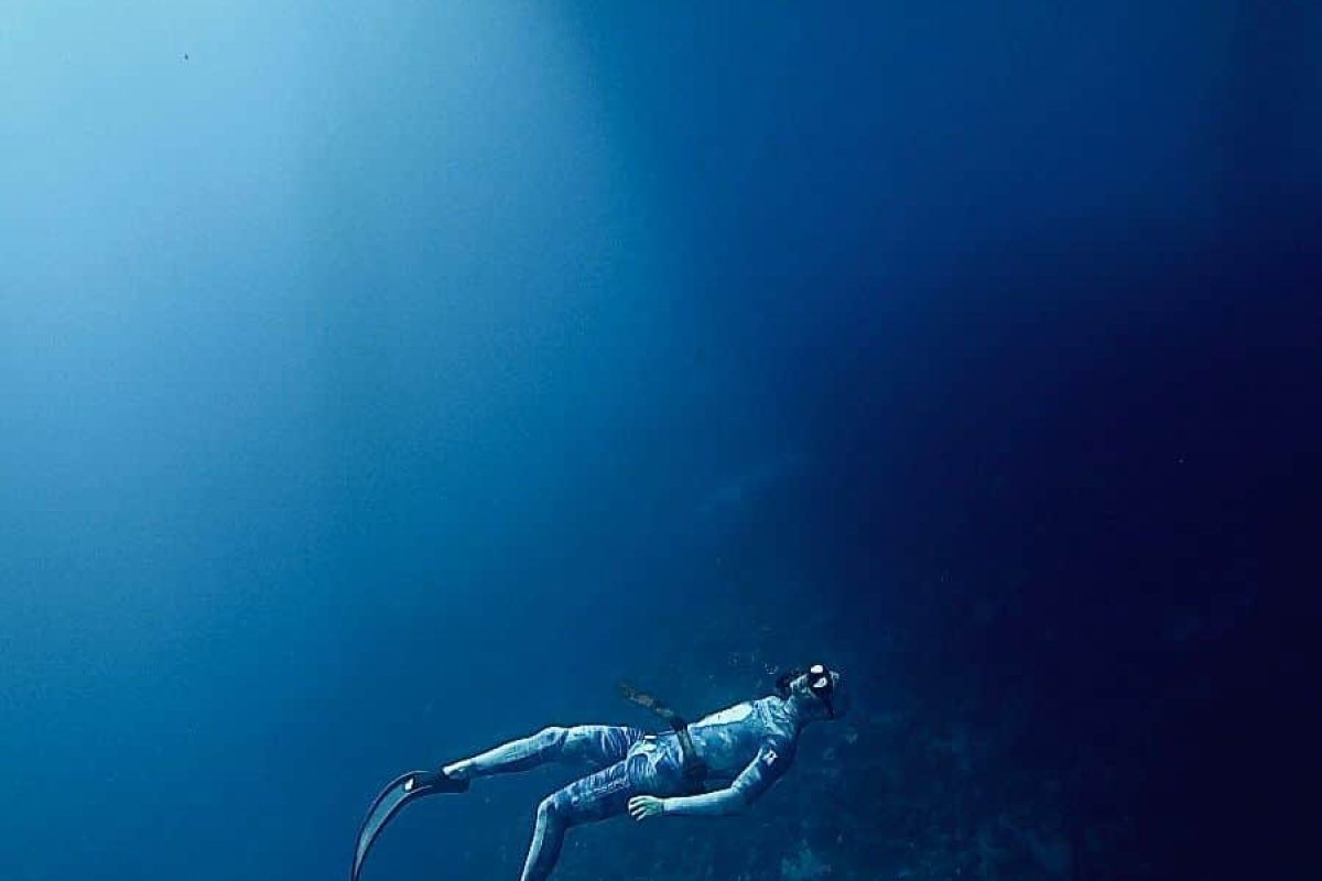 Experimenta el Curso de Buceo Libre para Principiantes (Bonaire): bucear bajo el agua con un traje de neopreno y aletas, rodeado por el océano azul profundo con la luz del sol brillando y el lecho marino visible abajo.