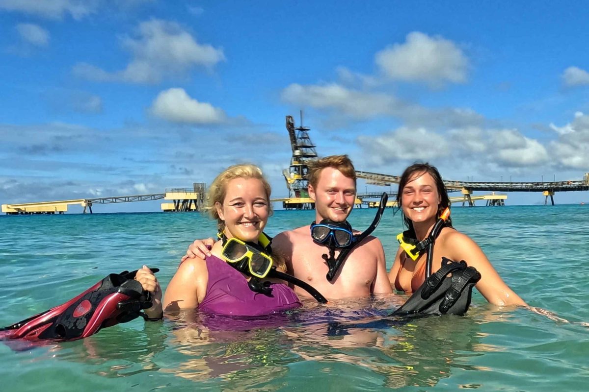 Tres personas con equipo de esnórquel sonríen mientras están en aguas claras y azules en un muelle industrial bajo un cielo soleado y parcialmente nublado durante el Tour Privado de Esnórquel (Bonaire).
