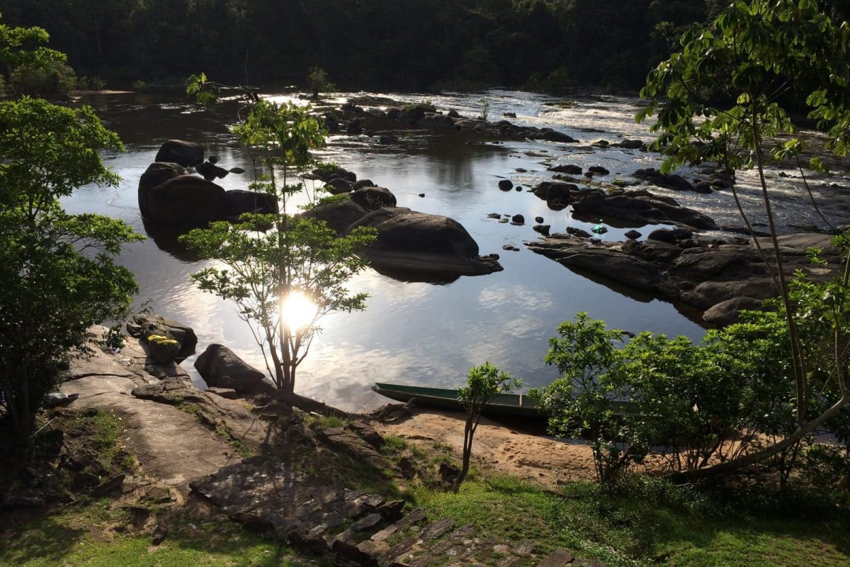 Ervaar de 4 Dagen Raleighvallen Voltzberg (Suriname): peddel langs rustige riviertaferelen met grote rotsen en zachte stroomversnellingen, weelderig groene bomen en zonlicht dat op het water glinstert terwijl de kano's langs de oever rusten.