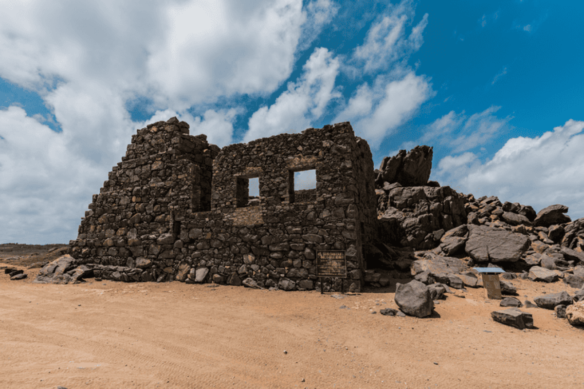 The Mangel Halto & Baby Beach Off-Road Safari (Aruba) shows a stone ruin on sandy soil under a partly cloudy sky, surrounded by scattered rocks and boulders, with a small sign near the building.
