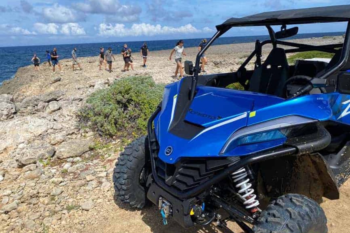 Un vehículo Off-Road Buggy Tour (Curazao) está aparcado en un terreno rocoso junto al mar, con personas caminando de fondo bajo un cielo parcialmente nublado.