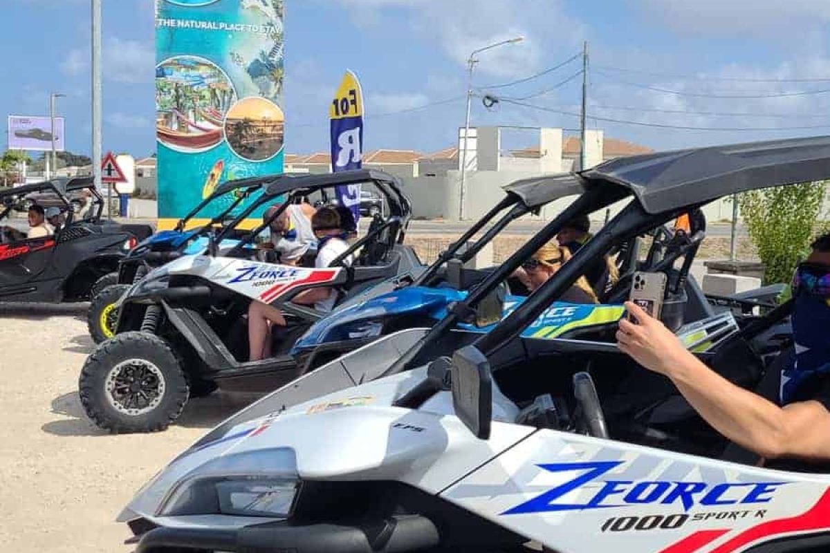 Un grupo se prepara con gafas de sol y cascos para el Tour de Buggy Todoterreno (Curazao), listo para explorar bajo cielos soleados, con un cartel colorido y edificios de fondo.