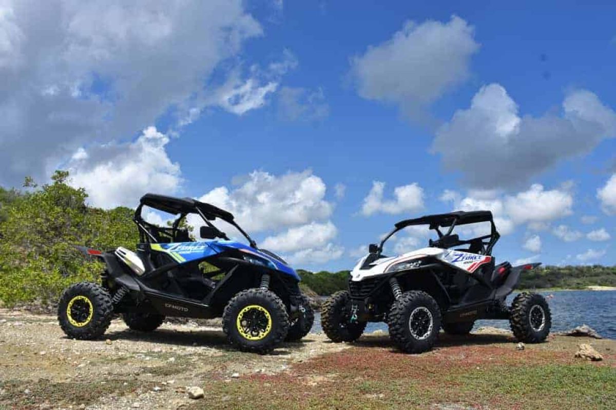 Dos vehículos Off-Road Buggy Tour (Curazao) – uno azul y uno blanco – están aparcados en terreno rocoso cerca del agua, bajo un cielo azul con nubes dispersas, con poca vegetación de fondo.