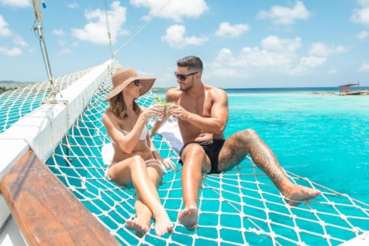 Een man en vrouw relaxen in een hangmat op de boeg van een Zeil en snorkel cruise (Aruba) boot over helder turquoise water, glimlachen en houden drankjes vast in zwemkleding en zonnebrillen onder een zonnige blauwe hemel.